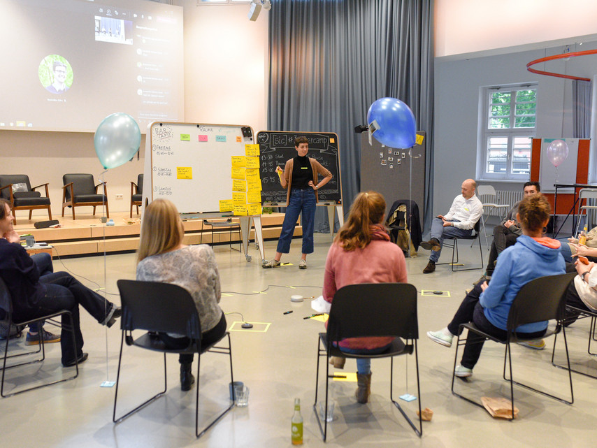 Man sieht eine Gruppe von Menschen in einem halben Stuhlkreis sitzen und auf die Rednerin vor dem Flipchart schauen.